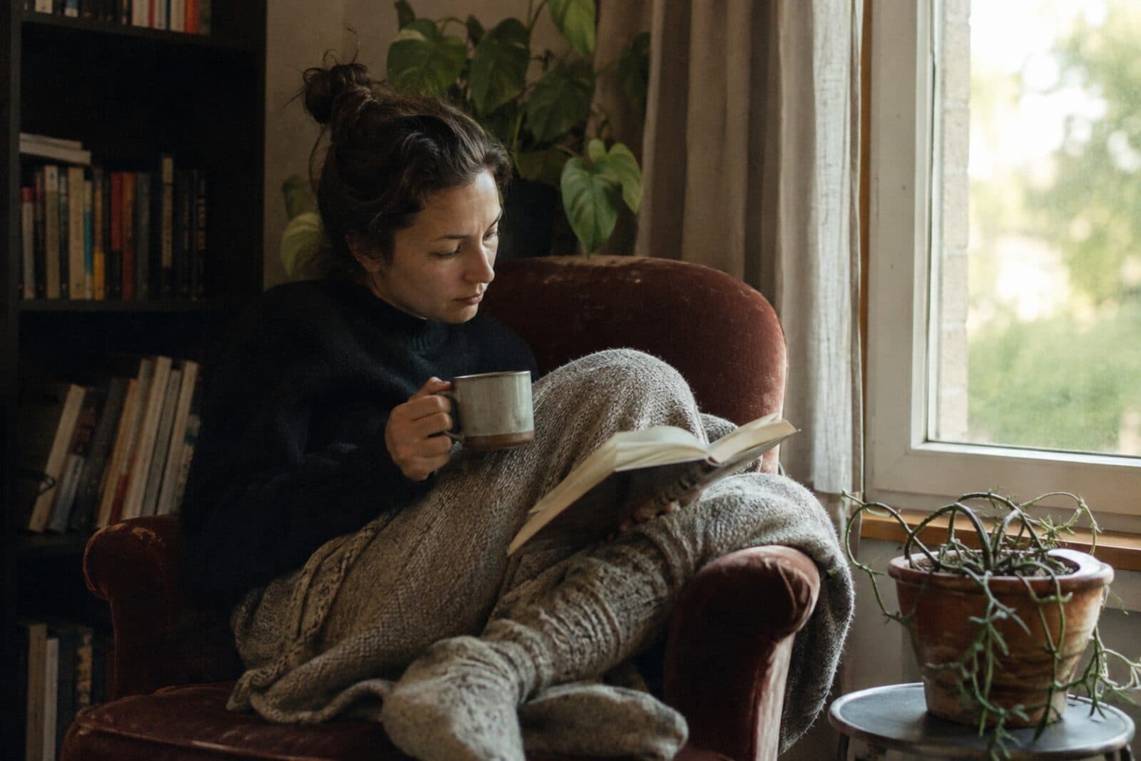 Eine Frau liest gemütlich im Sessel am Fenster, mit Kaffee, ohne Handy